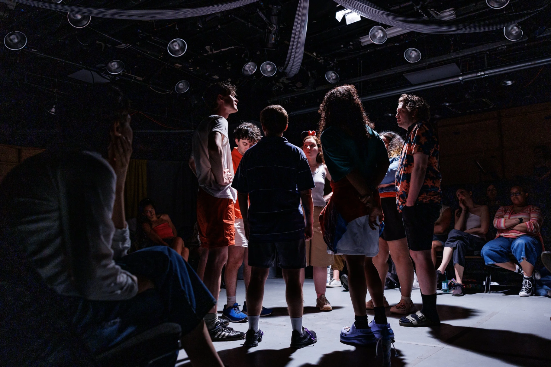 Students standing in a circle inside a small black-box theater under stage lights.